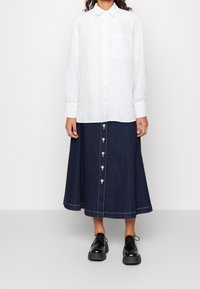 Biała lniana koszula zapinana na guziki z pojedynczą kieszenią na piersi, zestawiona z dżinsową spódnicą midi z guzikami z przodu i białymi akcentami stębnowania.