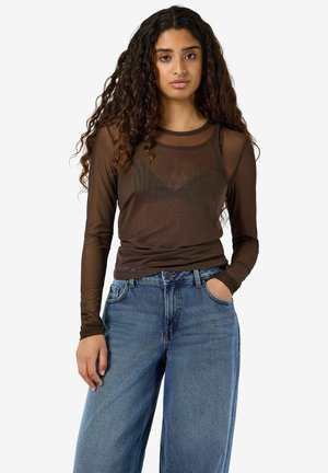 Femme aux longs cheveux bouclés portant un haut marron transparent à manches longues par-dessus un soutien-gorge marron et un jean bleu taille haute à jambes larges.