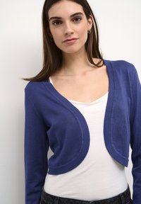 Blue cropped cardigan with rounded edges, made of soft knit fabric. Worn over a white tank top and paired with dark jeans.