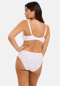 Femme aux longs cheveux foncés portant un soutien-gorge blanc et une culotte assortie avec des motifs de feuilles rose clair, se tenant avec la main sur la hanche, de dos.