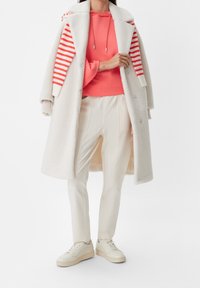 Biała puchowa kurtka z paskami w kolorze czerwono-białym, koralowy hoodie, kremowe spodnie i beżowe sneakersy. Gładkie tekstury i warstwowy design.