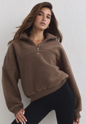 Joven mujer con cabello largo que lleva un suéter marrón de forro polar con cremallera y leggings negros, de pie frente a una pared clara con textura.