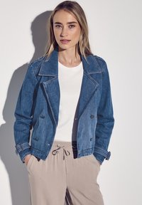 Jeansjacka i medelblått med spetsig krage, dubbelknäppta knappar och justerbara ärmar, kombinerad med en vit topp och beige byxor.