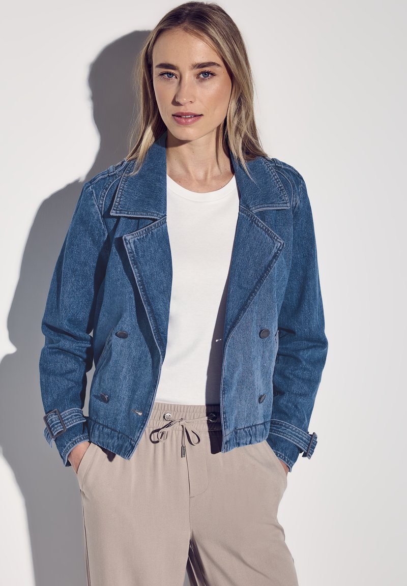 Jeansjacka i medelblått med spetsig krage, dubbelknäppta knappar och justerbara ärmar, kombinerad med en vit topp och beige byxor.