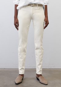 Jeans ajustados color crema con una textura suave y costuras sutiles, que presentan un cinturón con una hebilla metálica cuadrada y perneras acampanadas.