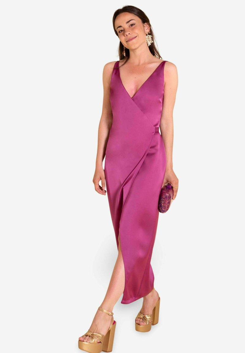 Vestido de satén fucsia con escote en V, diseño envolvente y abertura hasta el muslo. Combinado con tacones de plataforma dorados y un clutch texturizado púrpura.