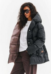 Kurtka puchowa z błyszczącym czarnym wykończeniem i kontrastującym matowym brązowym wnętrzem, z dużym kapturem i detalami w postaci zatrzasków.
