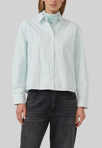 Vrouw in een lichtblauwe en witte verticaal gestreepte blouse over een lichtblauwe coltrui, gecombineerd met zwart vervaagde jeans.