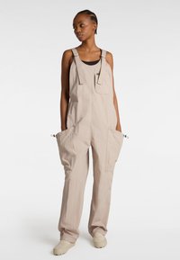 Beige Overall aus leichtem Stoff, mit großen Vordertaschen, verstellbaren Trägern und elastischen Bündchen an den Knöcheln. Lässiges Design.