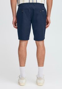 Shorts en coton bleu marine avec une coupe ajustée, dotés de poches arrière fermées par des boutons et d'une texture douce. Portés avec des chaussettes blanches et des baskets.