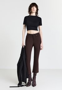 Top corto nero, jeans fitted marroni, giacca in suede nera appoggiata sul braccio e stivali con tacco in stampa leopardata. Sfondo semplice.