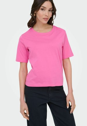 Woman wearing a bright pink short-sleeve cotton t-shirt and black pants, posing against a plain white background.
