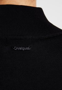 Närbild av svart ribbad tyg med hög krage och liten svart plast Desigual-logotyp sydd nära halsringningen.