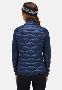Marineblauwe gewatteerde jas met een getailleerd ontwerp. Beschikt over een gladde textuur en een hoge kraag. Elastische manchetten en een zwarte hoofdband met witte tekst.