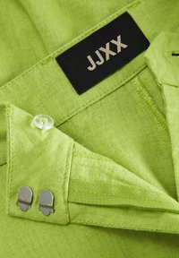 Zeleni materijal za odjeću s glatkom teksturom. Crna etiketa s natpisom "JJXX," prozirni gumb i metalni detalji na ovratniku.