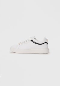 White sneakers with a textured finish, black accent stripe, gold eyelets, and logo detail. Features a thick white sole and a rounded toe.