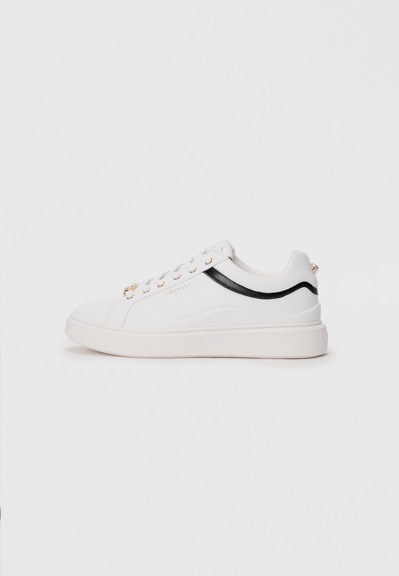White sneakers with a textured finish, black accent stripe, gold eyelets, and logo detail. Features a thick white sole and a rounded toe.