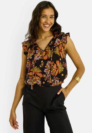 Femme souriante portant un chemisier noir sans manches avec un motif floral coloré et un pantalon noir, posant avec une main dans la poche.