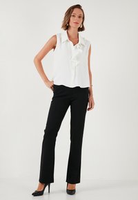 White sleeveless blouse with ruffled front detail, paired with black fitted pants and black heels. Smooth fabric and tailored fit.