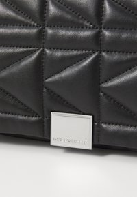 Juodas siuvinėtas odinis krepšys, turintis geometrinį raštą, su sidabriniu stačiakampiu logotipo plokšteliu, ant kurio išgraviruota "KARL LAGERFELD."