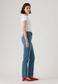 Λευκό κοντομάνικο μπλουζάκι συνδυασμένο με μπλε κανονικά τζιν. Το μοντέλο φοράει κόκκινες παπούτσια με γυαλιστερή δερμάτινη επιφάνεια. Ο απλός φόντος αναδεικνύει το σύνολο.