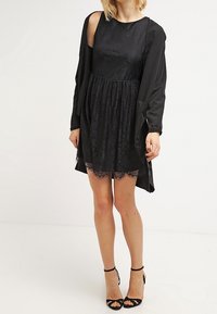 Black dress with lace accents, sleeveless design, elastic waist, and flowy overlay. Styled with black strappy heels.