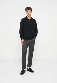 Zwart gebreid polo sweater met drie knopen, gecombineerd met grijs geruite broek en zwarte instappers met een lichte glans.
