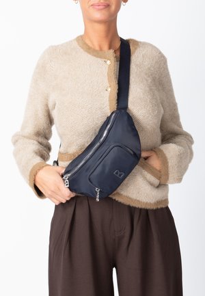 Donna con cardigan beige peloso e pantaloni marroni, con borsa a tracolla nera tenuta in vita, davanti a uno sfondo bianco.