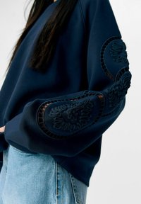 Marineblauwe sweatshirt met ingewikkelde geborduurde accenten op de mouwen, met een relaxte pasvorm en een zachte textuur, gecombineerd met lichte denim.