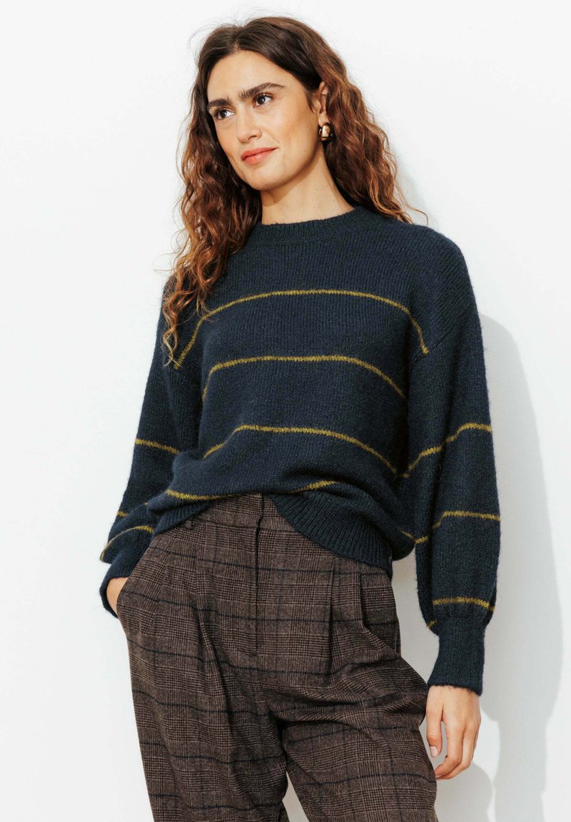 Pull rayé marine en maille douce avec des lignes jaunes, présentant une coupe décontractée et un col rond, associé à un pantalon taille haute marron à carreaux.