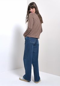 Vrouw met lang bruin haar, gekleed in een bruine lange mouw top, wijde blauwe jeans en witte sneakers, staand en over haar schouder kijkend.