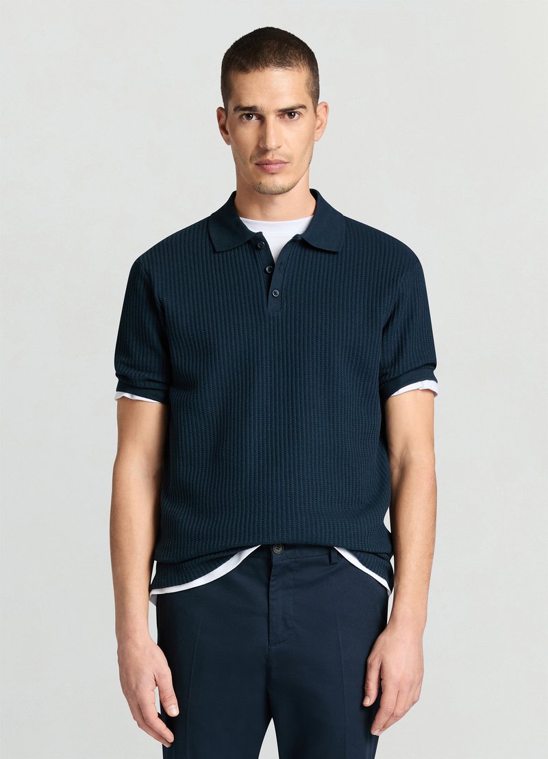 Polo bleu marine côtelé à manches courtes, avec col et patte à trois boutons. Porté par-dessus un t-shirt blanc.
