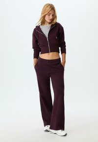 Felpa corta bordeaux con zip, maglietta grigia sotto e pantaloni abbinati; tessuto morbido, vestibilità comoda e tasche laterali; abbinata a sneakers bianche.