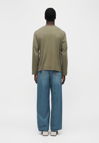 Άτομο που φοράει ελιά πράσινο μακρυμάνικο πουκάμισο, χαλαρό μπλε τζιν και λευκά παπούτσια με τετράγωνη μύτη, στέκεται γυρισμένο προς τα πίσω σε απλό φόντο.