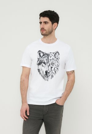 Férfi fehér pólóban, amelynek mintája félrealisztikus, fél geometrikus farkasarc, egy kézzel a zsebében áll, egyszínű háttér előtt.