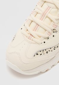 Sneaker de culoare crem, cu piele texturată, accente punctate și șireturi subțiri în dungi roz și alb, având o talpă groasă și suportivă.