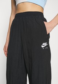 Černé kalhoty Nike Air s texturovaným povrchem, elastickým páskem a postranními kapsami, s bílým logem na levé noze.