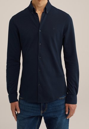 Mand iført en mørk navy knaplukket langærmet skjorte med krave, kombineret med blå jeans mod en ensfarvet baggrund.