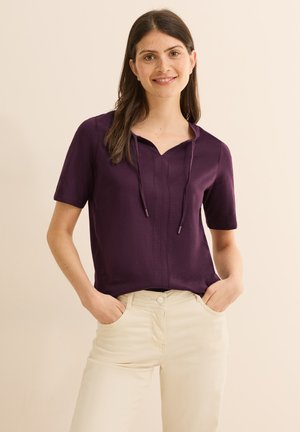 Camisa morada de manga corta y escote en V con cordones, que presenta una textura suave y sutiles detalles frontales, combinada con pantalones beige claros.