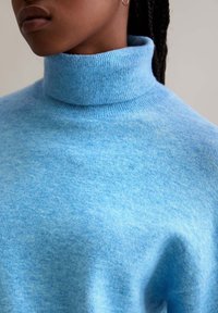 Helles bläuliches Rollkragenpullover aus weichem, strukturiertem Stoff. Hat einen hohen Kragen und gerippte Manschetten. Glatte Optik mit einem leicht melierten Muster.