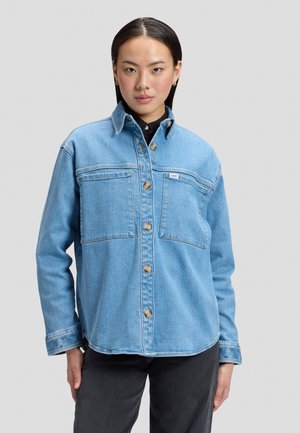 Femme portant une veste en denim bleu clair avec fermeture à boutons et grandes poches poitrine, debout devant un fond uni.