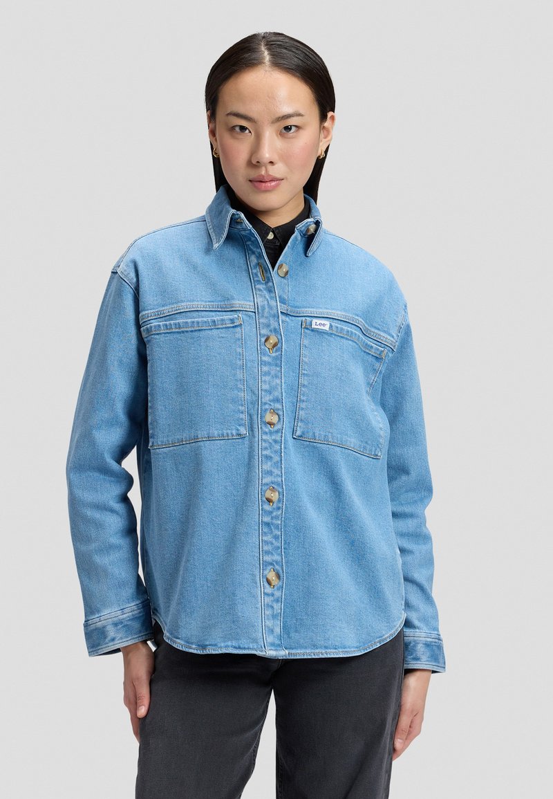 Femme portant une veste en denim bleu clair avec fermeture à boutons et grandes poches poitrine, debout devant un fond uni.