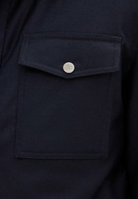 Camicia blu navy con tessuto testurizzato, dotata di una tasca sul petto con chiusura a scatto in metallo argentato e cuciture a contrasto.