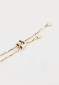 Bracciale in oro con due catene, caratterizzato da una barra centrale con marchio in rilievo. Piccole decorazioni rotonde su entrambe le estremità aggiungono un tocco di dettaglio.