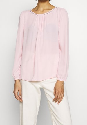 Vrouw die een lichtroze blouse met lange mouwen en geplooide voorkant draagt, gecombineerd met een witte broek, tegen een effen achtergrond.