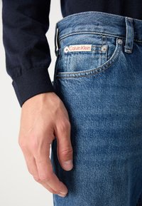 Niebieskie dżinsy z delikatnym przetarciem, z czerwonym metkiem Calvin Klein na przedniej kieszeni i srebrnym guzikiem. Zbliżenie na rękę na dżinsach.