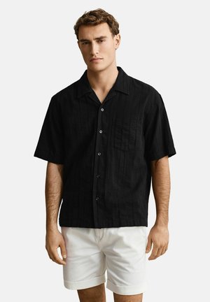 Mann trägt ein kurzärmliges schwarzes Hemd mit Knöpfen und weiße hochgekrempelte Shorts, steht vor einem einfarbigen Hintergrund.