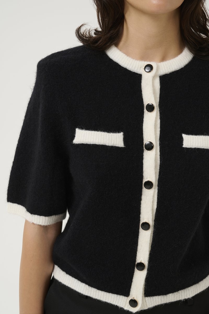 Kurze schwarze Strickjacke aus strukturiertem Stoff, mit weißem Besatz und Taschen, ausgestattet mit großen schwarzen Knöpfen auf der Vorderseite.