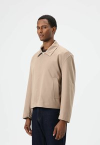 Beige cropped jasje met een puntkraag, zijzak en knoopsluiting. Gemaakt van gladde stof, met lange mouwen en een ontspannen pasvorm.