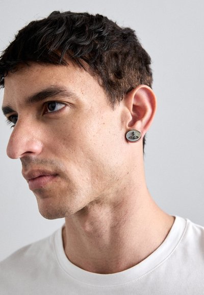 Oval metallørering med teksturert overflate, med et distinkt designelement, vist på en modell med kort hår og hvit skjorte.